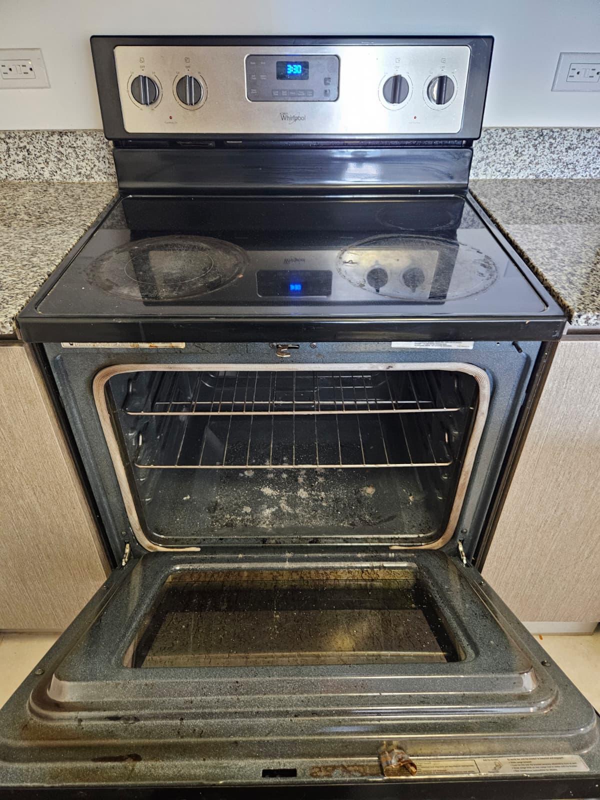 An open Whirlpool electric range: glass cooktop with burnt rings and crumbs, oven interior with baked-on residue on the floor and walls, discolored racks, and a dirty inner door